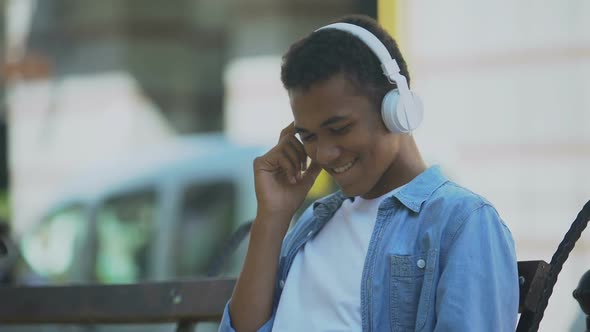 Positive Young Man Listening to Music in Headphones and Dancing Sitting on Bench