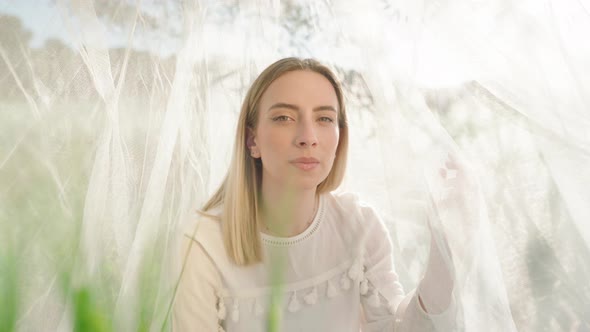 Pretty model woman posing into camera sitting on grass field surrounded by mosquito net during sunli