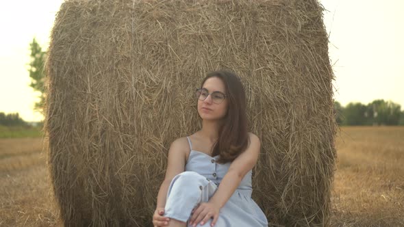 A Young Woman Sits Near a Haystack in a Field