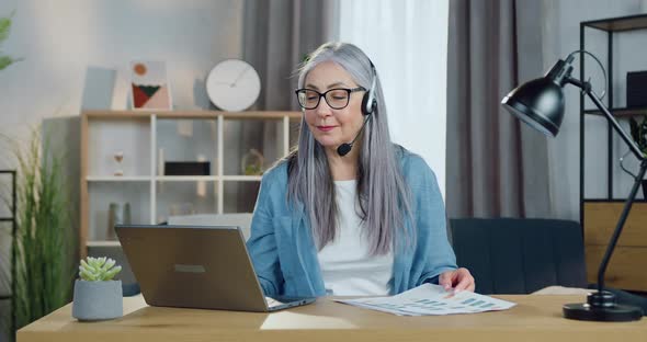 Senior Woman in Headphones Holding Online Meeting on Computer with Coworkers from Home office