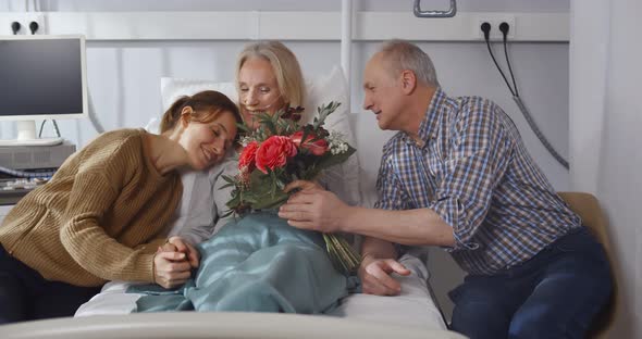 Happy Senior Husband and Young Daughter Visiting and Cheering Woman Lying in Bed at Hospital Ward