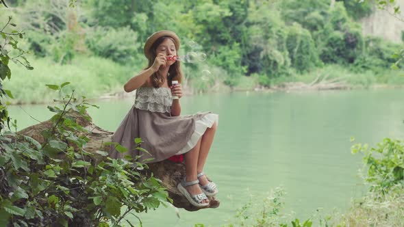 Pretty little girl sitting on the old log and blowing a soap bubbles. Summertime holidays