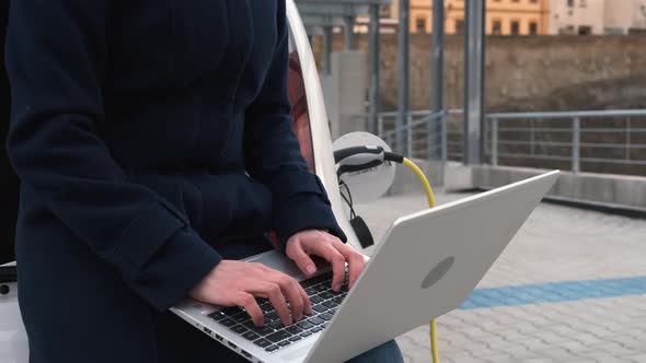 A Woman Sits on the Trunk of an Electric Car with a Laptop and Waits for the Car to Charge in the