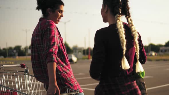 Camera Moves Around Two Attractive Stylish Girls Standing By the Shopping Cart on Parking and