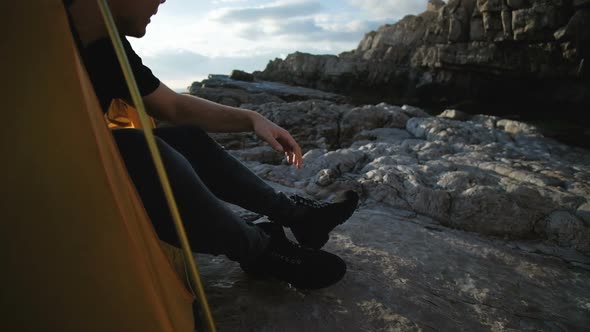 Young man in a black T-shirt is resting in a tent on the rocks by the sea