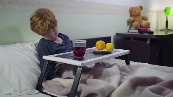 Sad Caucasian Redhead Boy Lying in Bed and Drinking Hot Fruit Tea. Portrait of Sick Child Staying