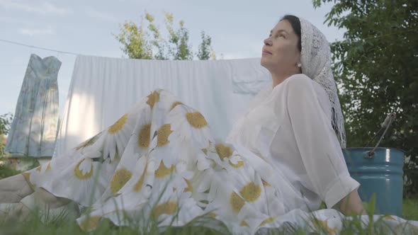 Cute Senior Woman with a White Shawl on Her Head Resting in the Garden Sitting on the Grass