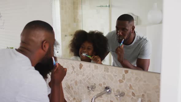 African american daughter and her father looking in mirror brushing teeth together in bathroom