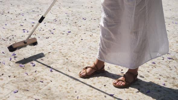 High Angle View of Young Woman in White Dress Sweeping with Broom Outdoors in Sunlight