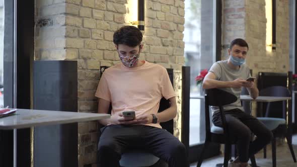 Two Young Caucasian Men in Face Masks Sitting in Cafe on Covid19 Pandemic Waiting for Order