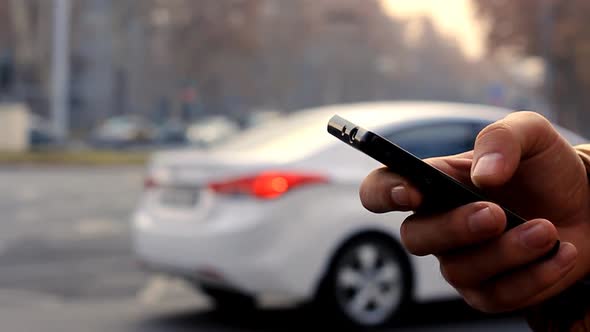 A Man's Hand Uses Black Smartphone on the Background Cityscape with Cars