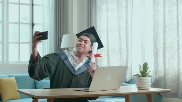 Asian Man Selfie While Wearing A Graduation Gown And Cap, Hand Holding University Certificate