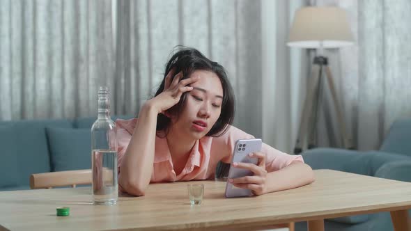 Drunk, Depressed Asian Woman Drinking Vodka From Glass During Using Smartphone At Home