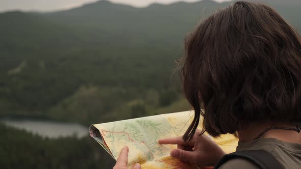 A young girl on a trip stands with a paper map against the background of mountains and a lake