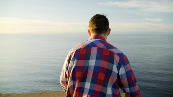 Young man walking towards the edge of a cliff, looking at sunset view