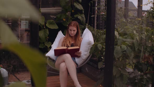 Teenage Girl Sitting On Garden Chair With Pillows And Reading