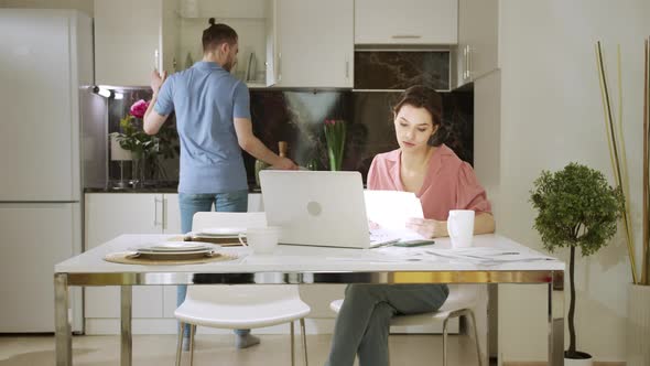 An Adult Woman is Sitting and Working at Home While Her Husband is Doing Household Chores