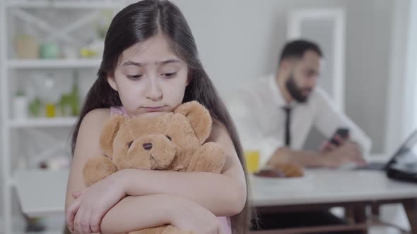 Portrait of Upset Middle Eastern Girl Hugging Teddy Bear and Looking at Camera with Blurred Father