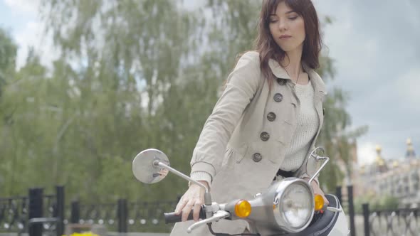 Confident Brunette Woman Coming To Motorcycle and Sitting on Scooter. Portrait of Elegant Young