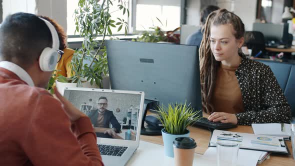 Young Man Making Online Video Call with Laptop While Women Colleagues Using Computers in Office