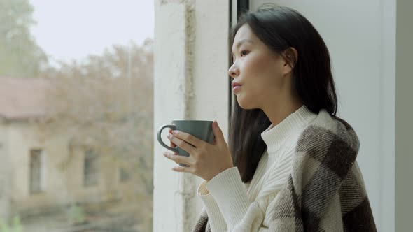 Young Sad Asian Woman Drinks Coffee or Tea and Looks Out the Window Covered with a Blanket