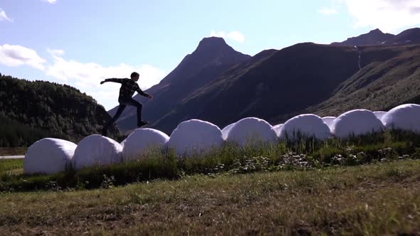 Slow motion shot of man jumping over bales of hay on a beautiful day with mountain background.