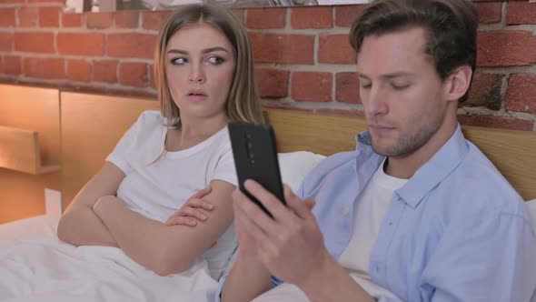 Upset Young Couple Using Smartphone in Bed