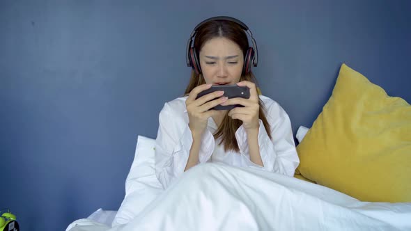 Happy teen woman wearing headsets playing online game with a smart phone on a bed