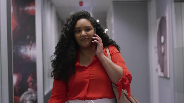 Smiling biracial businesswoman talking on smartphone walking down corridor in modern office