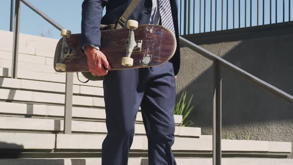 Midsection of caucasian businessman walking downstairs, holding skateboard on sunny day