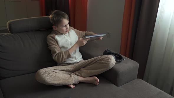 Teen Boy Reads a Book Sitting on the Couch. He Puts the Book Aside and Takes the Headphones To