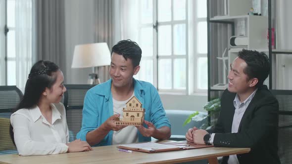 Asian Couple With A House Purchase Contract Paper Receives The House Model From A Real Estate Agent