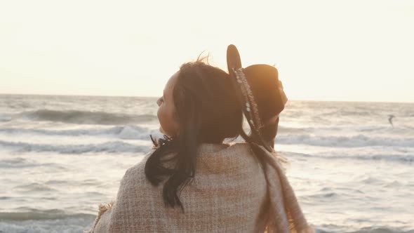 Portrait of Romantic Brunette Woman in Hat and Coat Walking on the Beach in Autumn at Sunset or