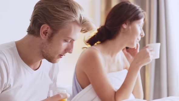 Attractive Young Caucasian Pair Sitting in Bed in the Morning Drinking Juice and Coffee Reading