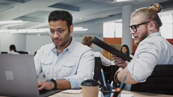 Indian Male Working at Notebook on Business Project Sitting at Table in Office Caucasian Colleague