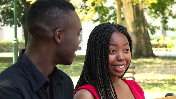 A Black Man and a Black Woman Sit on a Bench and Talk in a Park on a Sunny Day - Closeup