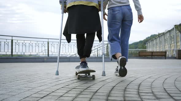 Young Man with One Leg on Crutches Riding on Skateboard Walking with His Girlfriend