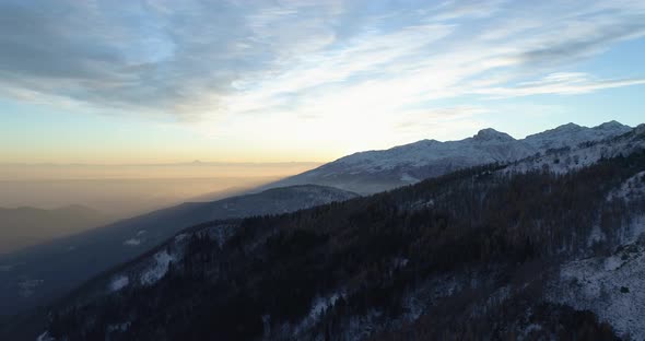 Backward Aerial Top View Over Winter Snowy Mountain and Woods Forest at Sunset or Sunrise