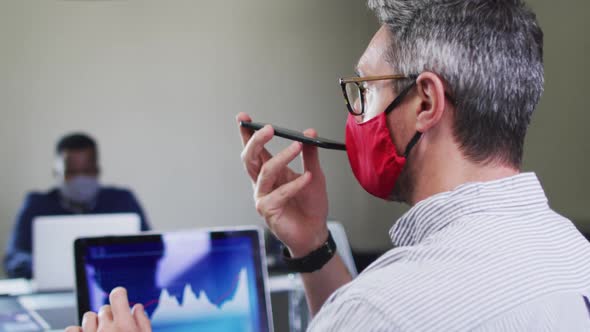 Caucasian man wearing face mask talking on smartphone while sitting on his desk at modern office