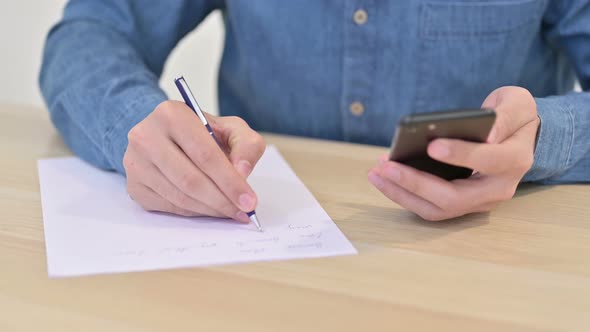 Close Up of Man Writing on Paper Using Smartphone 