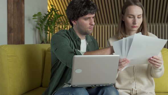 Young Couple Sitting on Yellow Sofa at Home and Managing Budget Using Laptop