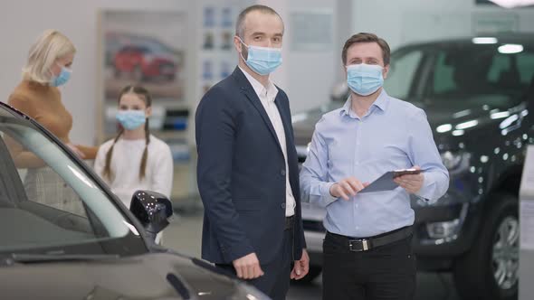 Cheerful Men in Covid19 Face Masks Looking at Camera Showing Thumbs Up As Blurred Woman and Girl