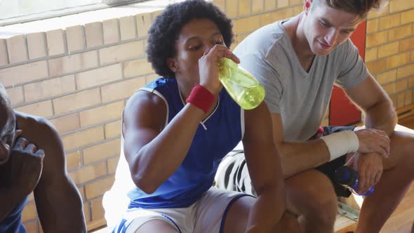 Diverse male basketball team and coach resting and drinking water after match