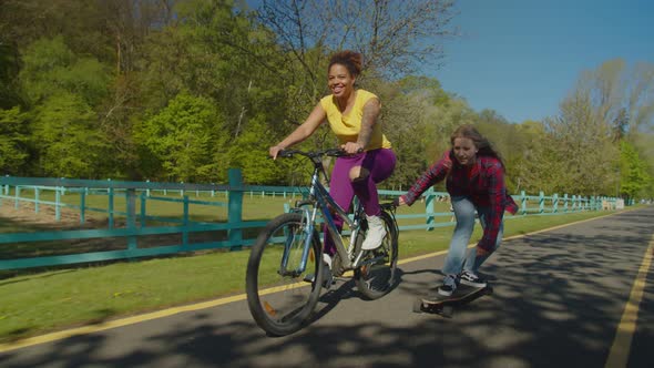 Lovely Black Woman Cyclist Speeding Up Female Skater During Skateboarding Outdoors