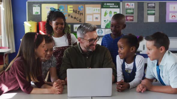 Diverse male teacher and group of schoolchildren looking at laptop