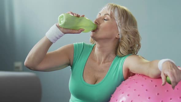 Senior woman leaning on fitness ball, drinking water after workout, hydration