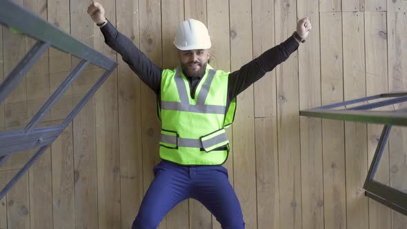 Smiling Builder in the Helmet and Green Jacket Lying on the Floor Showing Victory Gestures