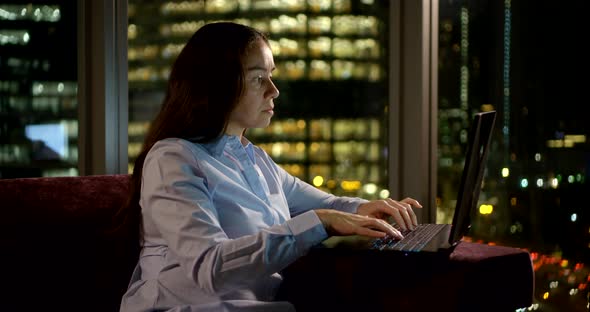 a Middle-aged Woman with Long Dark Hair Is Working on a Computer By a Large Window. Outside the