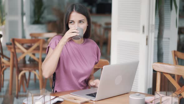 Beautiful Young Woman Freelancer Drinks Coffee and Uses Laptop for Work in Cafe