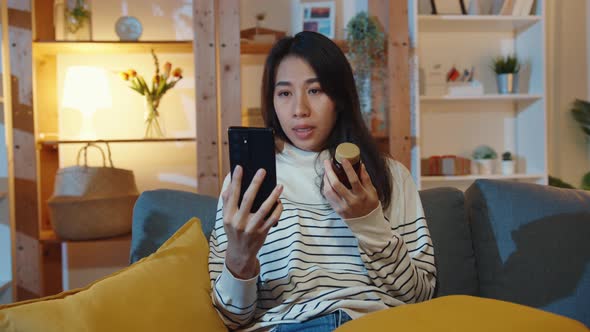 Sick young Asian lady hold medicine sit on couch video call with phone consult with doctor.